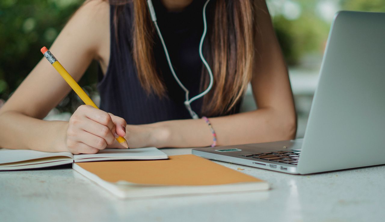 Crop anonymous young female freelancer with earbuds writing notes with pencil in notebook while sitting at table with laptop in contemporary cafe