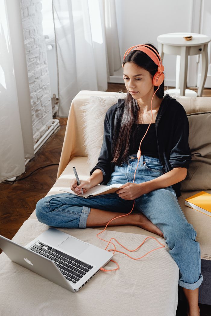 Teen girl sitting on a sofa, wearing headphones, using a laptop and taking notes indoors.